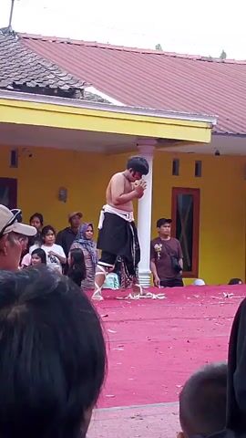 The video shows a performance taking place outdoors, possibly in a village or residential area. In the foreground, the backs of several audience members are visible, with one person in a beige baseball cap in profile on the left. The stage is covered with a bright red carpet, and scattered leaves or petals are on the carpet. In the center of the stage, a performer, a young male, is standing. He is shirtless, wearing a black sarong or cloth wrapped around his waist, and white leg wraps. He is holding a small, light-colored object in his hands and appears to be bringing it to his face, possibly to smell or eat it. His face is painted with dark markings around his eyes. Behind the performer, a yellow building with a tiled roof and visible support columns is present. Several spectators are gathered, including women and children, some seated and some standing, watching the performance. To the right of the performer, another young male spectator is visible. The overall atmosphere seems to be that of a community event or cultural performance.