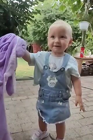 A young child with blonde hair and wearing denim overalls over a light blue t-shirt, is holding a purple stuffed animal in their right hand. The child is standing on a brick-paved patio. In the background, there is a lush green garden with trees and bushes. Further back, there is a wooden table with a red thermos and a blue bucket on it. Clothes are hanging on a clothesline in the upper right portion of the frame. The child turns and walks away from the camera, towards the wooden table, with the stuffed animal still in hand.