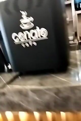 The video shows a close-up of a counter in what appears to be a cafe or coffee shop. There are several coffee-making appliances, including a coffee grinder with a clear hopper full of beans and a black base. To the left, a blue machine is partially visible, its purpose unclear from this shot. To the right, a large black machine with a white logo that appears to say "CO" is present. The counter itself has a polished, marbled surface. In the background, a glass display case and shelves can be seen, suggesting the area is a service counter. The lighting is bright and appears to be from overhead fixtures.