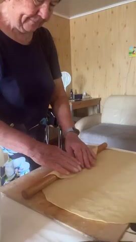 A person is seen rolling out dough on a wooden board. The person is wearing a dark blue t-shirt and a floral apron. Their hands are actively engaged in pressing and moving a rolling pin across the pale yellow dough, which is spread thinly. The scene appears to be indoors, with wood-paneled walls in the background. A white couch is visible behind the person, and a small picture hangs on the wall.