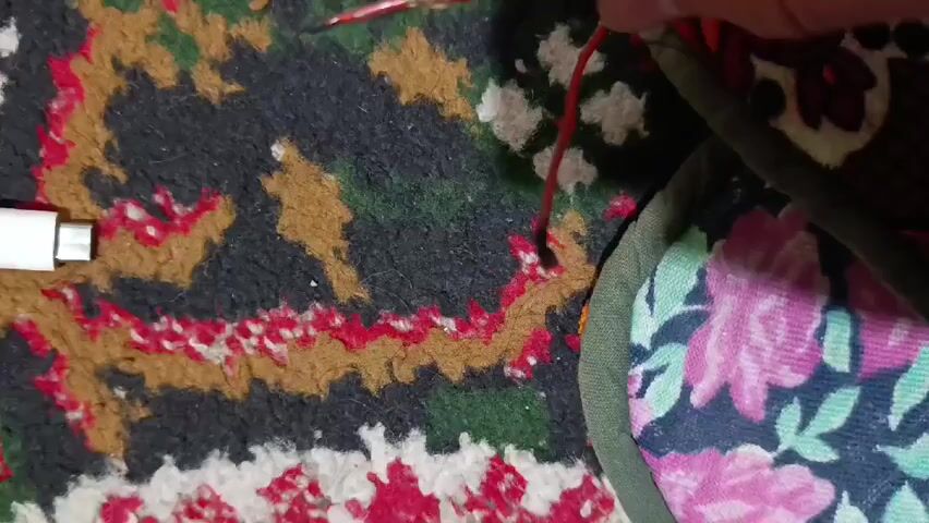 A person holds two exposed red wires that touch, causing a spark. The sparks start out purple and then turn orange. The wires are held over a rug with a brown, red, and green pattern, next to a pair of slippers with a floral pattern. A white USB charging block sits on the rug to the left.