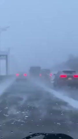 The view from inside a vehicle shows cars driving on a highway in heavy snow. The road is covered in a layer of snow, and visibility is significantly reduced due to the heavy snowfall. The tail lights of the vehicles ahead are visible through the snow and fog. There are visible streaks on the windshield from the wipers. Overhead, there are indistinct street lights.