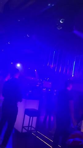 The video shows a dimly lit nightclub with vibrant purple and pink lights. In the foreground, a person in a black shirt is standing on the left, facing away from the camera. To the right, another person in a black shirt is also standing, facing away. In the middle ground, there's a brightly lit bar with a stool in front of it. A person is partially visible behind the bar, and another person is sitting on the stool. The background is dark with some illuminated structures and more colored lights. The atmosphere appears to be energetic, with people likely enjoying themselves in a social setting.