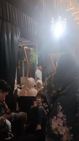 The video shows a dimly lit indoor gathering, possibly a wedding reception or party, with a strong overhead light source causing a hazy effect. Several people are seated and standing, many wearing headscarves and modest clothing. In the foreground, a young boy sits on a dark chair, looking at his phone. A man in a white shirt stands near a decorative archway with artificial greenery. The space is decorated with hanging white fabric strands and ornate chandeliers. The overall atmosphere appears festive, with guests engaged in conversation and observation.