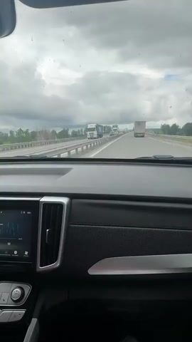 The video shows the interior of a car as it travels on a highway. The view is primarily from the front windshield, with occasional shots from the side window and the rearview mirror. The sky is overcast with heavy clouds. Several trucks and cars are visible on the highway, with traffic appearing to be moderately heavy. The car's dashboard and interior are visible in the foreground, including a dashboard screen and air vents. The landscape outside the car consists of fields and trees, with a guardrail running alongside the highway. The perspective shifts to show the side of the car, including the door panel and the side-view mirror, which reflects the passing scenery.
