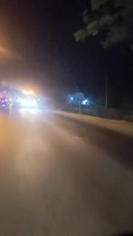 The video shows a nighttime scene on a road. It appears to be a moving vehicle's perspective, with the road and passing vehicles in motion. Several vehicles are visible, including large trucks and smaller cars. Many of the vehicles have bright lights, including headlights, taillights, and illuminated signs. Some of the lights are white, while others are yellow or blue. The overall lighting conditions are dim, with the vehicle lights creating significant glare and streaks of light. Trees and streetlights are visible along the side of the road in some shots. The road surface appears to be wet or reflective, contributing to the light effects. The video is brief and shows a continuous, albeit blurry, sequence of driving at night.