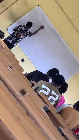 The camera moves from low to high to showcase a classroom scene with students. Initially, a person stands at a whiteboard, writing. The camera pans across rows of seated students. Most are facing forward, presumably paying attention.  Several individuals are visible wearing shirts with numbers and logos. The classroom is equipped with wooden desks and yellow walls, and a window is partially visible. The general setting suggests an academic environment.