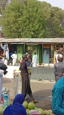 The video shows a busy roadside scene with various individuals. Many of the people are wearing traditional clothing. Some are standing, some are sitting, and some are walking. One man is on a phone call. There are stalls and structures along the side of the road. There are also boxes, bags, and produce visible. In the background are trees. It appears to be a daytime scene.