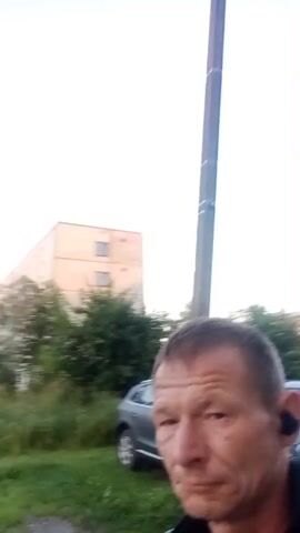 A man with short brown hair and a slight stubble is in the foreground of the video. He is wearing a black and yellow athletic shirt and earbuds. He is looking directly at the camera. In the background, there is a large, light-colored apartment building with several floors and windows. Trees and grass are visible in front of the building. There is a dark-colored SUV parked to the right of the man. The sky is clear and light blue, suggesting it is daytime. The video appears to be taken outdoors, possibly in a residential area.