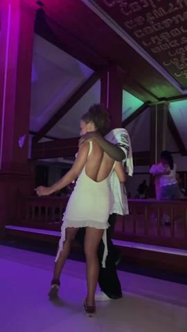 A couple slow dances at a nighttime event indoors. The setting features prominent wooden pillars and railings, with dynamic lighting that shifts between purple, green, and red hues. The woman, with curly hair, wears a backless white dress with a ruffled or tattered hem. The man, with a beard, wears a white jersey-style t-shirt, possibly with "JAY" visible, and dark pants. His arm is around her shoulder as they sway gently.

In the background, other individuals are visible, some dancing or socializing on an upper level or behind railings. The overall atmosphere seems relaxed and enjoyable. A notable feature of the location is the decorative ceiling or wall adorned with intricate script, possibly Thai or Khmer, which aligns with the provided location context of Muang Pattaya, Thailand. The indoor architecture with its high ceiling and wooden elements suggests a resort, hotel lobby, or event hall.