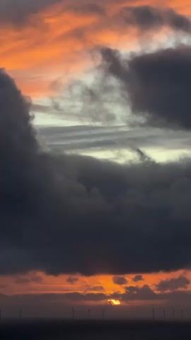 The video captures a dramatic sunset or sunrise over the ocean near Fécamp, France. The sky is filled with a striking contrast of dark, heavy grey clouds and vibrant streaks of orange and red, illuminated by the low sun. The sun itself is partially visible, a glowing orange orb peeking through a break in the clouds just above the horizon. Below the dramatic sky, a line of offshore wind turbines stands silhouetted against the luminous backdrop, stretching across the dark expanse of the water. The clouds appear to be in motion, adding a dynamic element to the serene yet powerful scene.