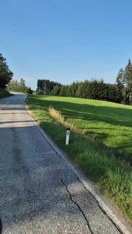 The video shows a road with green grass and trees on the side. The road is asphalt and has white lines. The sky is blue. The camera is moving, suggesting it is from a vehicle.