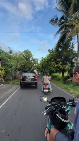A man is driving a motor scooter down a paved road. The camera is mounted on the scooter facing forward and angled slightly towards the driver’s face. The driver is wearing sunglasses, a sleeveless blue shirt and shorts, and flip-flops. The scooter has a design on it and a helmet is strapped to the front. There is a car driving in front of the scooter and another woman riding a scooter further ahead. The road is surrounded by green trees. There are other scooters parked on the side of the road. The sky is blue with some white clouds.