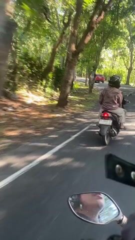 A first-person perspective video shows a motorcyclist riding on a paved road lined with trees. The road has a white painted line on the right side. In front of the camera, a motorcycle is visible, with its mirror reflecting the rider's face. The rider in front is wearing a brown jacket and a black helmet, riding a scooter with a passenger behind them. In the distance, other vehicles and people can be seen. The environment appears to be tropical or sub-tropical, with lush green foliage. The video captures the movement of the motorcycle, indicated by motion blur and the changing perspective of the surroundings.
