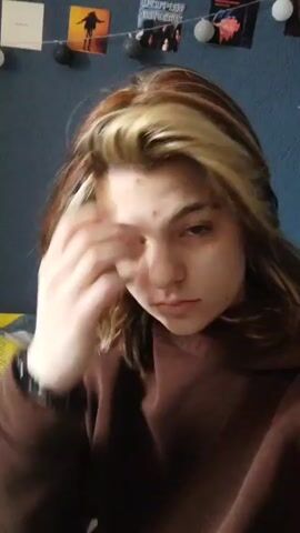 A young woman with fair skin and light brown hair with blonde highlights sits on a bed, wearing a brown sweatshirt and black pants. She touches her hair and looks down. The bed has a colorful patterned blanket with yellow, gray, and white geometric shapes. A fluffy white cat sits next to her on the bed. The wall behind the bed is dark blue, and decorative string lights are visible along the top of the wall.