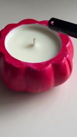 A person is lighting a candle in a bright pink, pumpkin-shaped container. The candlewick is white and the flame is small and yellow. The container is sitting on a white surface. The background is out of focus and appears to be a white surface. The video is shot from a close-up angle.