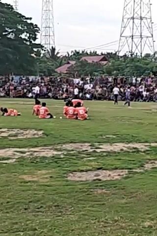 The video shows an outdoor soccer game with a large crowd watching from the sidelines. The field is grassy with a goal at one end. There are several players on the field, some wearing orange jerseys and others wearing blue jerseys. Spectators are seated and standing around the perimeter of the field, some are very close to the action. In the background, there are trees, houses, and power lines. The sky is overcast.