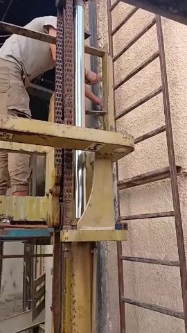 A man stands on a yellow elevated platform, reaching towards a shiny metal cylinder attached to a vertical yellow structure. He's wearing a light-colored shirt and cargo pants, with a silver bracelet on his wrist. A heavy chain is positioned to the left of the cylinder. A metal ladder is affixed to the wall, which appears to be textured and painted a light beige color. A black pipe runs overhead in the upper portion of the frame.