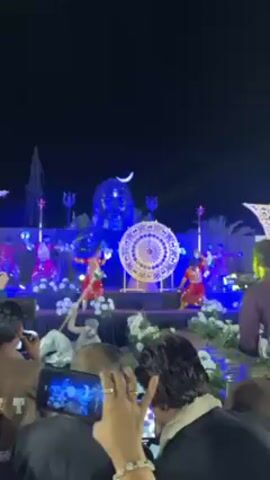 A low-angle shot depicts several people on a stage at night, illuminated by bright blue stage lighting. In the background stands a large, illuminated sculpture of what appears to be the head of a deity with a crescent moon above its head. In front of the sculpture, a large, decorative, round object dominates the center of the stage. Several people in traditional red costumes dance with sticks, their movements synchronized. 
Below the stage, a crowd watches, many holding up their phones to record the performance. The immediate foreground shows the backs of peoples' heads and shoulders, with some holding their phones aloft.