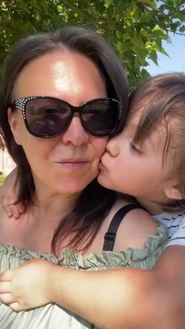 This video features a close-up shot of a woman and a child. The woman, wearing sunglasses and a green off-the-shoulder top, is being hugged and kissed by the child. The child, with brown hair and a striped shirt, has their arm around the woman's neck. The background suggests an outdoor setting with green foliage and a blue sky, indicating daytime. The woman is smiling, and the child appears happy and affectionate. There is no visible text in the video. The context provided places the location in Altopascio, Italy.