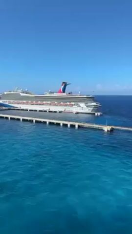 The video shows a cruise ship docked at a pier. The ship is white with red and blue accents, and it has a large, distinctive funnel on top. The pier is made of concrete and extends out into the ocean. The water is a vibrant turquoise color, and the sky is clear and blue. A small island can be seen across the water. The camera appears to be moving slightly, providing a panning view of the scene.
