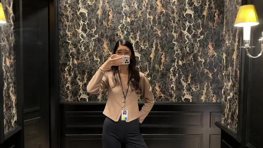 A woman is standing in a bathroom taking a selfie in the mirror. The bathroom is decorated with dark wallpaper with a marble pattern. The lower half of the wall is a black panel. Two sconces with yellow lamps are on either side of the mirror. The woman is wearing a light brown blazer, dark pants, and an ID badge. She is holding her phone up to take the photo.