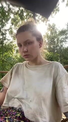 A woman sits in a chair outdoors, partially obscured by a large floral-patterned fabric. She wears a loose, cream-colored t-shirt and has her hair pulled back. Tattoos are visible on her arms. The background consists of trees and foliage, with sunlight filtering through the leaves. The camera is positioned low, looking up at the woman. The fabric, which appears to be part of her clothing or a blanket, is moved during the clip, revealing her legs and feet.