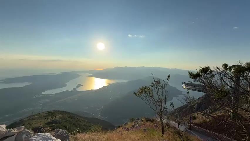 The video shows a panoramic view from a mountaintop at sunset. The sun is low in the sky, casting a golden reflection on the water below. Mountains frame the scene, and a body of water, likely a bay or fjord, stretches into the distance. A small town or village is visible nestled along the coastline. In the foreground, dry grass and a few scattered flowers are visible, along with rocks. To the right, a tree with sparse leaves and a railing or pathway suggest a viewing area or a hiking trail. A modern structure, possibly a restaurant or viewing platform, with a distinctive rounded edge, juts out over the landscape on the right side of the frame. The sky is clear blue with a hint of orange and pink near the horizon, indicating the sunset. No people are visible in the video.