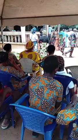 The video shows a gathering of people under a white tent. Most of them are sitting on blue plastic chairs and facing away from the camera. The people in the foreground are wearing shirts with a patterned design of orange and blue. One woman is wearing a yellow shirt. An older man with a bald head is sitting in the second row wearing a brown shirt. In the background, more people are standing outside the tent. A building with white pillars can be seen behind the people outside the tent. There is singing.