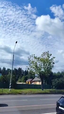The video shows a view from a vehicle moving along a road. The sky is blue with scattered white clouds. On the right side of the road, there is a row of trees and a house visible. A black car is driving on the road. A white vehicle, possibly a truck or van, is partially visible on the far right edge of the frame. The video pans slightly, showing a street lamp and a fence along the roadside.