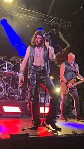 The video shows a male musician on a stage. He has long, dark, wavy hair and is wearing a black bandana, a black leather vest, and black leather pants. The vest is open, revealing his chest. The pants have a decorative scarf-like accessory hanging from the left hip, adorned with what appear to be skulls. He is also wearing a necklace with a cross pendant and several bracelets on his left wrist. He is standing with his left leg slightly forward, holding a microphone. A drum set is visible behind him to the left, and another person, also wearing a black vest, is partially visible to the right holding a guitar. The stage is lit with colorful lights, including blue, yellow, and red. There are speakers and other stage equipment visible. The background includes a dark curtain with a stylized logo.