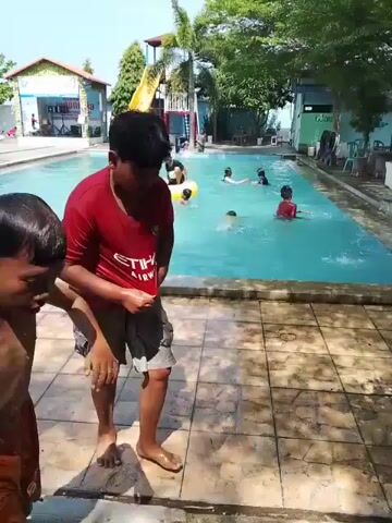 The video shows a sunny day at a swimming pool with several children and adults. In the first shot, a boy in a red shirt and shorts is standing near the edge of the pool, facing the camera. The camera then pans to show the entire pool area. There are people swimming and playing in the pool. The pool is surrounded by tiled decking and trees. There is a yellow water slide in the background, along with some buildings and a dolphin statue. The camera follows children as they run, swim, and play near the pool.