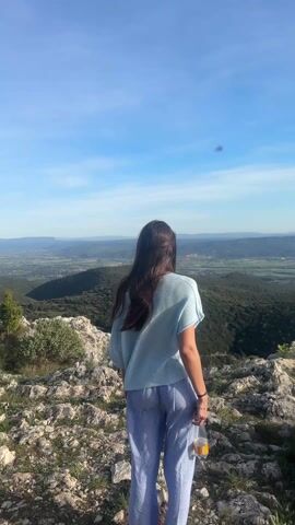 A woman is captured from behind, standing on a rocky outcrop overlooking a vast landscape. She has long, dark brown hair that falls past her shoulders and appears to be blowing slightly in the breeze. She is wearing a loose-fitting, light blue short-sleeved top and light blue and white vertically striped pants. In her right hand, she holds a clear plastic bottle, possibly containing water or a light-colored beverage, with an illegible label. A dark bracelet or band is visible on her left wrist.

The setting is a panoramic view from what appears to be a mountain or hilltop, likely in Bonnieux, France. The foreground consists of rough, light-colored rocks and sparse, low-lying green vegetation. Beyond this immediate area, rolling green hills and valleys stretch into the distance, dotted with trees and what might be small towns or agricultural fields. Further in the background, more distant mountains or higher hills frame the horizon. The sky above is a clear, bright blue with a few scattered, faint white clouds, indicating sunny weather. The overall impression is one of natural beauty and a peaceful, expansive vista. The time of day appears to be bright daytime, likely late morning or afternoon. No discernible text is clearly visible beyond the potential label on the bottle.