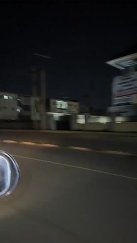 A blurry view of a dark street at night. Buildings and streetlights are visible in the background as the camera moves along the road.