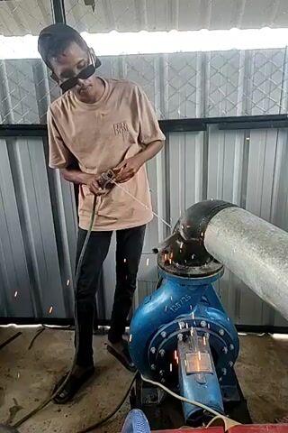 A young man wearing a baseball cap, sunglasses, and a tan t-shirt stands next to a large blue pump. He is holding a green hose connected to the pump and a white string. He appears to be operating the pump. Sparks are visible near the connection of the pump and a large pipe. In the background, there is a corrugated metal wall and a white roof. Another person is visible in the last frame, sitting and looking towards a large pipe. The environment appears to be outdoors, possibly a construction site or industrial area.