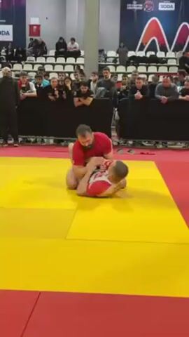 The video shows two men grappling on a yellow and red mat. One man is wearing a red shirt and the other is wearing a red and white shirt. The man in the red shirt has his legs wrapped around the other man's waist. They are both on the ground, facing each other. There is a crowd of people watching them. The scene is taking place indoors, likely in a large arena or gymnasium, as evidenced by the presence of bleachers, banners with logos, and an elevated platform separating the participants from the audience.