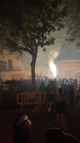 The video captures a nighttime outdoor event with a crowd of people gathered. A large tree with full foliage dominates the center-left of the frame, casting shadows. In the background, buildings with illuminated windows are visible. The primary focus of the scene is a spectacle of fire and light occurring in the distance, with bright orange and yellow flames erupting and sparks or fireworks being launched upwards. Several people in the foreground and midground are holding up smartphones, seemingly recording the event. They are standing on a paved area, possibly a street or square, with a yellow barrier in the middle ground separating the onlookers from the action. The atmosphere appears festive and energetic, with the bright lights of the display illuminating the smoke and haze. Some individuals are silhouetted against the light, while others are more clearly visible. The overall impression is of a public celebration involving pyrotechnics.