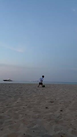 A young boy wearing a white and light blue striped t-shirt and black shorts is playing with a soccer ball on a sandy beach. The boy is kicking the ball with his right foot. In the background, there is a body of water and a large white boat. The sky is a light blue color with a few wispy clouds. The lighting suggests it is either early morning or late evening.