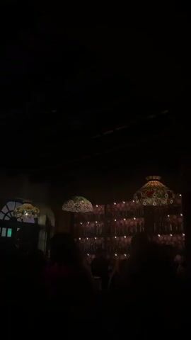 The video shows a dimly lit interior space, possibly a restaurant or bar, with several people silhouetted in the foreground. Ornate lamps with stained-glass shades hang from the ceiling. In the background, a wall is decorated with numerous small, glowing bottles or lights, creating a warm, inviting ambiance. The overall impression is one of a cozy, atmospheric evening setting.