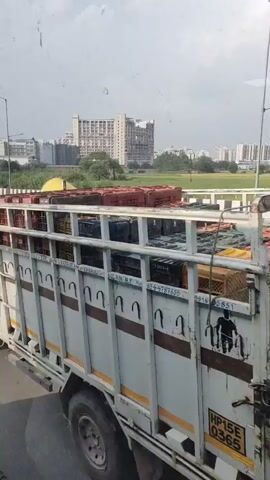 The video shows a white pickup truck driving on a road. The truck is carrying a load of red and yellow crates, some of which are stacked on top of the truck's canopy. In the background, there are buildings and trees. A motorcycle with two people on it is visible on an overpass to the left of the truck. The sky is cloudy. The truck has a license plate that reads "MP 15 E 6763". The scene appears to be in India.