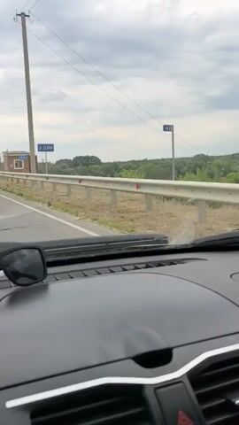 The video shows a view from inside a car driving on a road. A sign with blue text that appears to be in Cyrillic is visible on the side of the road, along with a small brick building and a fence. The car's dashboard and windshield wipers are also in view. The sky is cloudy.