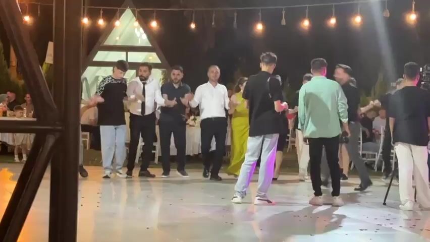The video shows a group of people dancing outdoors at night. The scene is illuminated by string lights and lights emanating from a triangular-shaped structure in the background. People are dressed in casual and formal attire, and some are holding hands while dancing in a circle. The dance floor is wide and appears to be made of a light-colored material, with some red confetti scattered on it. A few individuals are standing and observing the dancers, while others are actively participating in the dance. There are tables and chairs visible in the background, suggesting that this may be a wedding reception or a similar celebration. The overall atmosphere seems festive and lively.