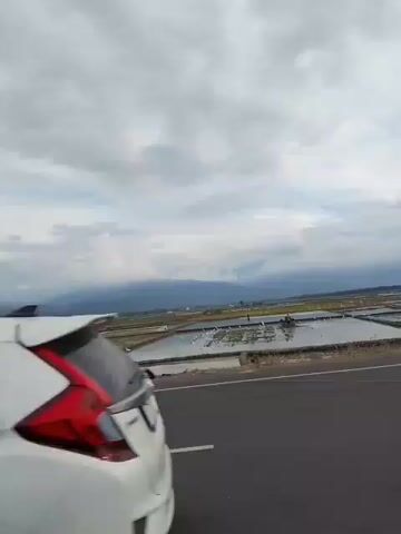 The video shows a rural landscape with a road in the foreground. Beyond the road are several flooded agricultural fields, likely rice paddies, with some dry earth visible between them. In the distance, mountains are shrouded in clouds. The sky is overcast with a mixture of gray and white clouds. A single person on a black scooter, wearing a helmet, rides from left to right across the frame. Several white birds are scattered around one of the flooded fields. The overall impression is of a vast, open, and somewhat overcast rural environment.