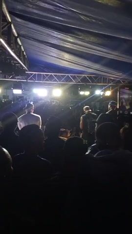 The video shows a nighttime outdoor concert with a live audience. A stage is set up under a large, dark tarp supported by metal scaffolding. Bright stage lights illuminate the performers and the crowd. Two male performers are visible on stage. One, wearing a white t-shirt and a baseball cap, stands with his back to the camera, facing the audience. The other performer, wearing a dark sleeveless shirt and a baseball cap, is positioned further upstage, holding a microphone and singing or speaking. Numerous audience members are in the foreground, mostly silhouetted against the stage lights, with many holding up their phones. The crowd appears dense and engaged with the performance. The overall atmosphere suggests a lively and energetic event.