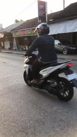 The video shows a side-view of two people riding motorcycles on a street. The person in the foreground, on a red and black scooter, is wearing a light blue helmet, a dark gray hooded sweatshirt, dark pants, and a purple backpack. They are riding with their right foot bare and resting on the scooter. The person in the background, on a white and black scooter, is wearing a dark jacket and a dark helmet. They are riding slightly ahead of the foreground scooter. The street appears to be paved and is lined with buildings on the right side, suggesting an urban or semi-urban environment. The lighting suggests it is either early morning or late afternoon, with the sun casting a warm glow. The overall scene depicts everyday street life.