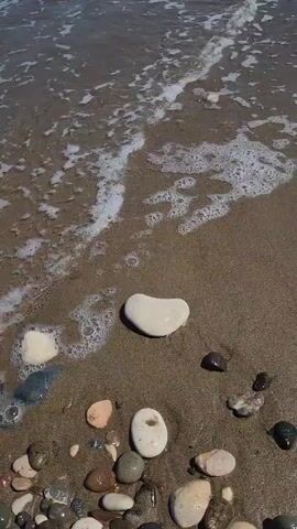 The video displays a static, high-angle shot of a beach with wet sand and various sizes of smooth, colorful pebbles and rocks, including a prominent heart-shaped white rock in the center. Gentle waves from the ocean, visible in the upper portion of the frame, roll in and recede, wetting the sand and rocks. The water is a dark, murky color with white foam. The sand is dark and wet, reflecting light in some areas. The rocks are smooth and varied in color, with some appearing dark, some light gray, and some white. The video is silent.