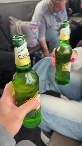 The video captures a lively scene inside a crowded passenger vehicle, likely a bus or train. In the foreground, a person's hand holds a green glass bottle of Harbin Beer, featuring visible Chinese characters "哈尔滨" and "HARBIN BEER" on its label. This hand clinks bottles with another person's hand, adorned with bright pink fingernails, a silver bracelet, and a smartwatch, also holding an identical Harbin Beer bottle.

The individual with pink nails, wearing a light grey long-sleeved top and blue jeans, then looks at a vibrant pink mobile phone, her expression appearing neutral or slightly pensive. A blue shoulder bag rests on her lap. In the background, several other passengers are visible, some engaging with their phones, others seated and looking around. The interior of the vehicle has dark seating, suggesting standard passenger transport.

Audible Russian dialogue provides crucial context for the activity. A male voice says, "Let's go, a video for arriving on the Chinese side, let's go!" The woman with the pink nails responds playfully, "Wait, I haven't had time yet!" followed by, "Alright, that's it, I didn't have time." This exchange, coupled with the Harbin beer (a Chinese brand) and the location context of Nizhneleninskoe, Russia (a border town), strongly indicates a celebratory moment marking an arrival or crossing into China. The scene appears to take place during the daytime under general indoor lighting. The atmosphere is relaxed and celebratory, with a touch of good-natured banter.
