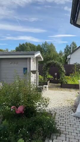 The video shows a backyard scene on a sunny day. In the foreground, on the left, is the side of a gray shed with a white trim. A broom leans against the shed. A pink flamingo lawn ornament is visible among some green plants. A hanging basket is suspended from the shed. The ground is paved with interlocking gray stones. In the middle ground, an archway covered in green vines stands over a path. There are raised garden beds on either side of the path. A white plastic stool and some black containers are on the paving stones near the shed. In the background, a brown wooden fence separates the yard from a wooded area with trees. A white house with a chimney is visible to the right. The sky is blue with white clouds.