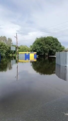The video shows a flooded street with standing water. In the background, there are several buildings and a large tree. A corrugated metal fence is partially submerged in the water. The sky is cloudy.