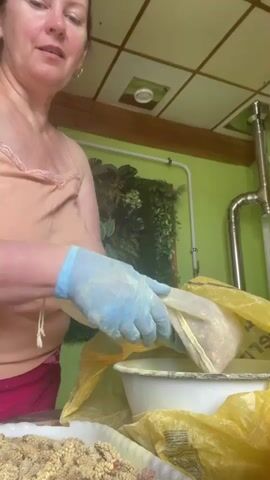 A woman with light skin and dark hair is putting a bag of ground material into a white bowl. She is wearing an orange tank top, red shorts, and blue latex gloves. The bowl is sitting inside a yellow trash bag. There is also a tray of the same ground material on a table in front of her. The room has green walls and a tiled ceiling. There is an indoor plant decoration in the background.